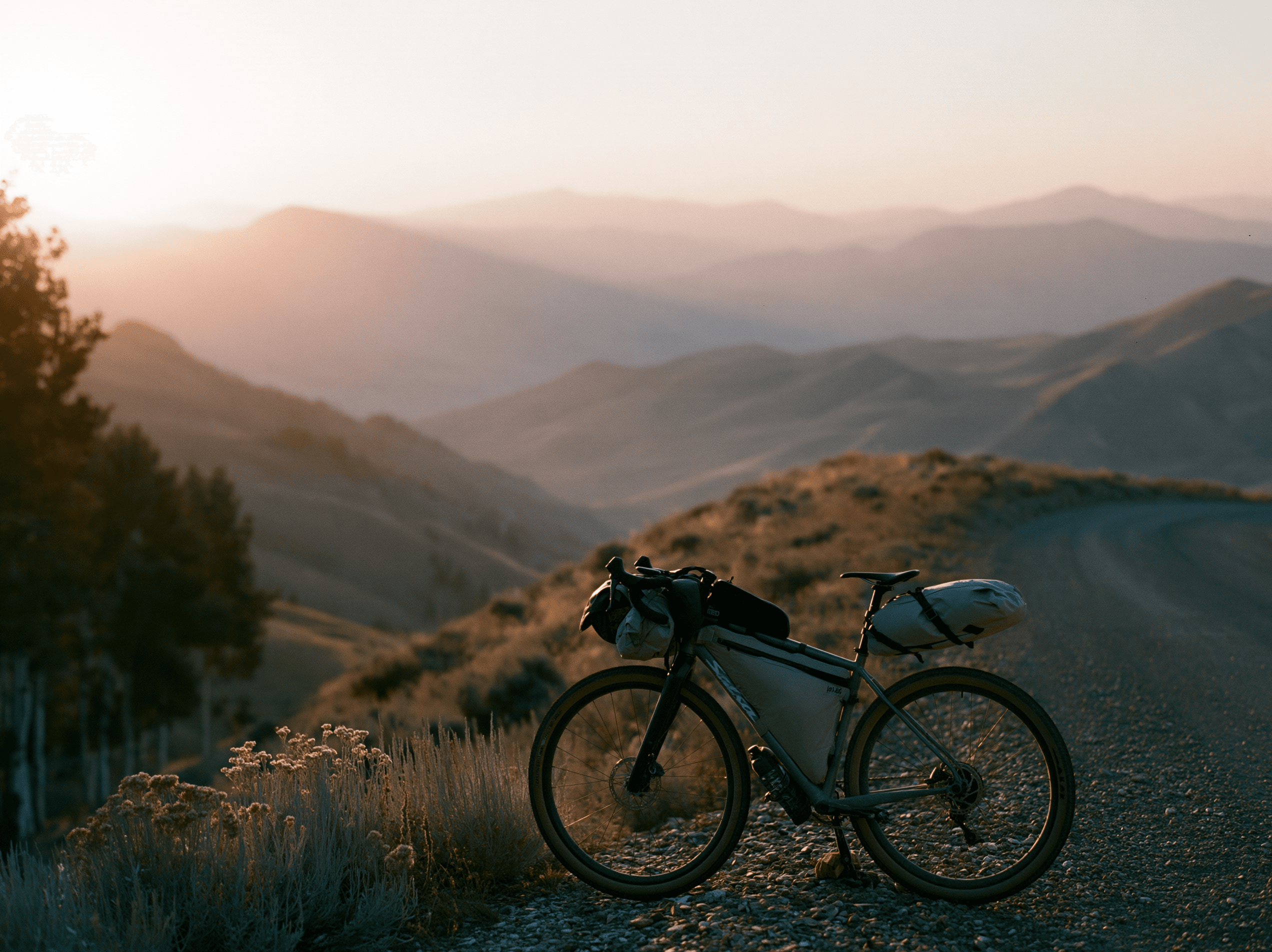 A loaded bikepacking bike pulled off a gravel ridge road at sunset, mountain ranges fading into the distance.