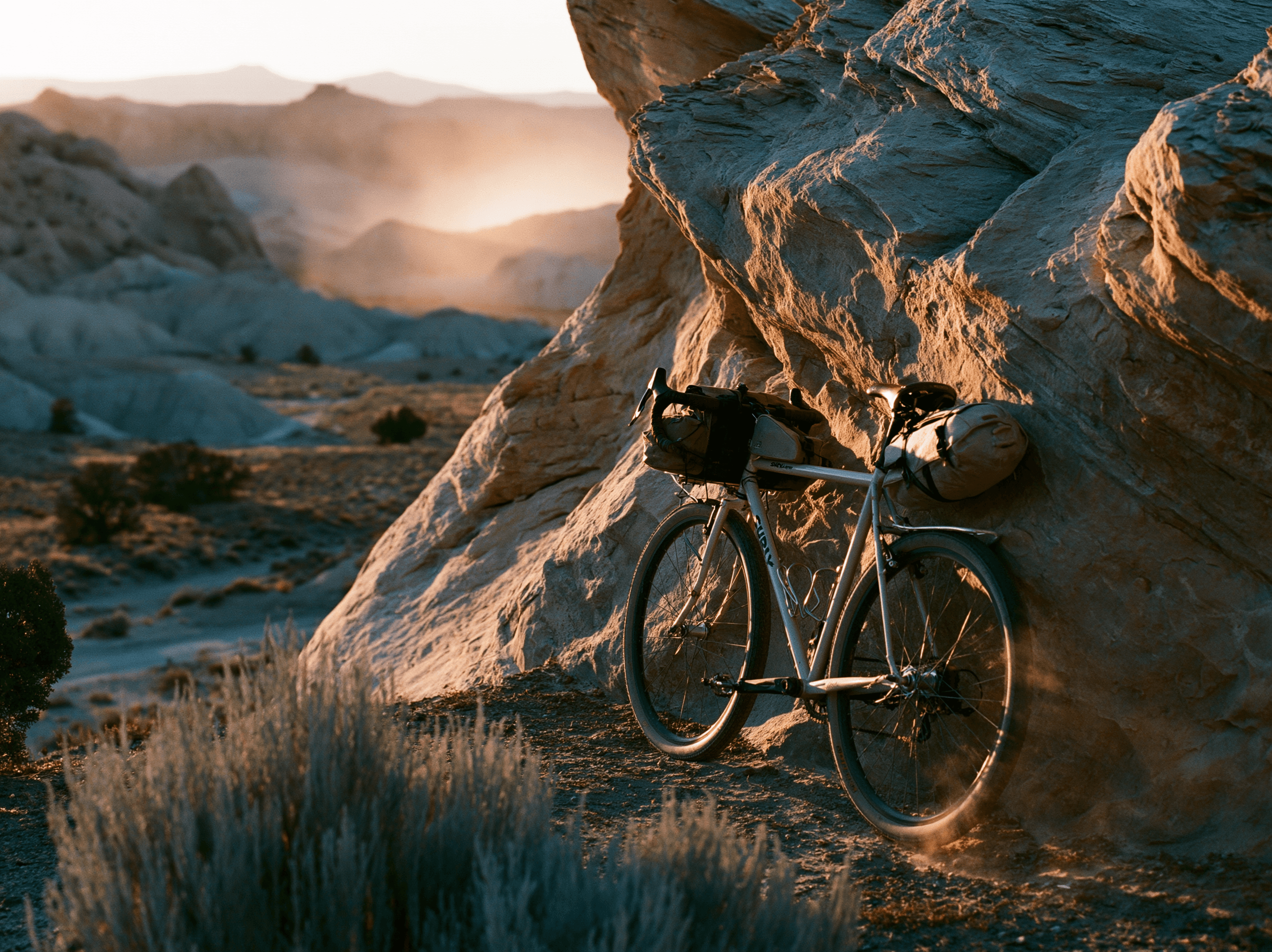 A loaded steel touring bike leaning against a sandstone wall in a desert canyon at sunset.