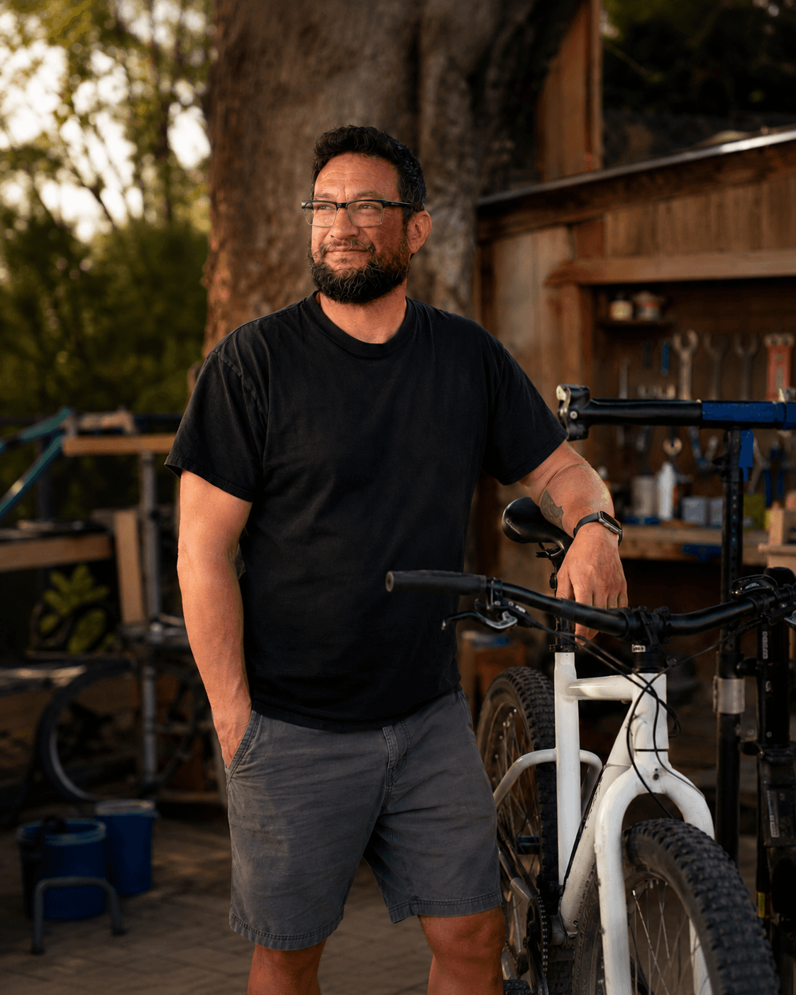 Seth Parsons standing beside a bike in a workstand at his outdoor workshop, warm sunset light.