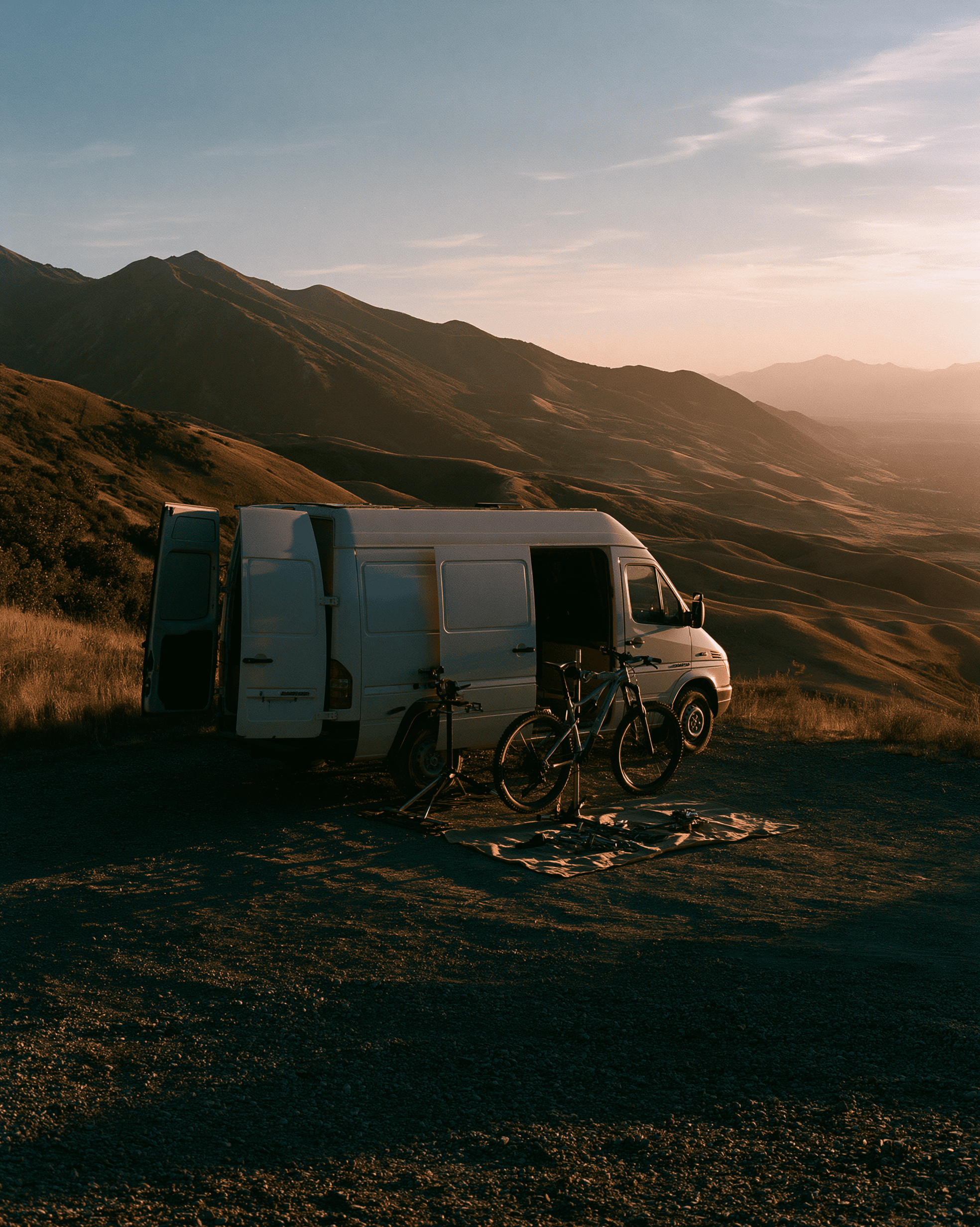 A white service van with its side door open and two bikes leaning against it, parked at a mountain overlook at sunset.